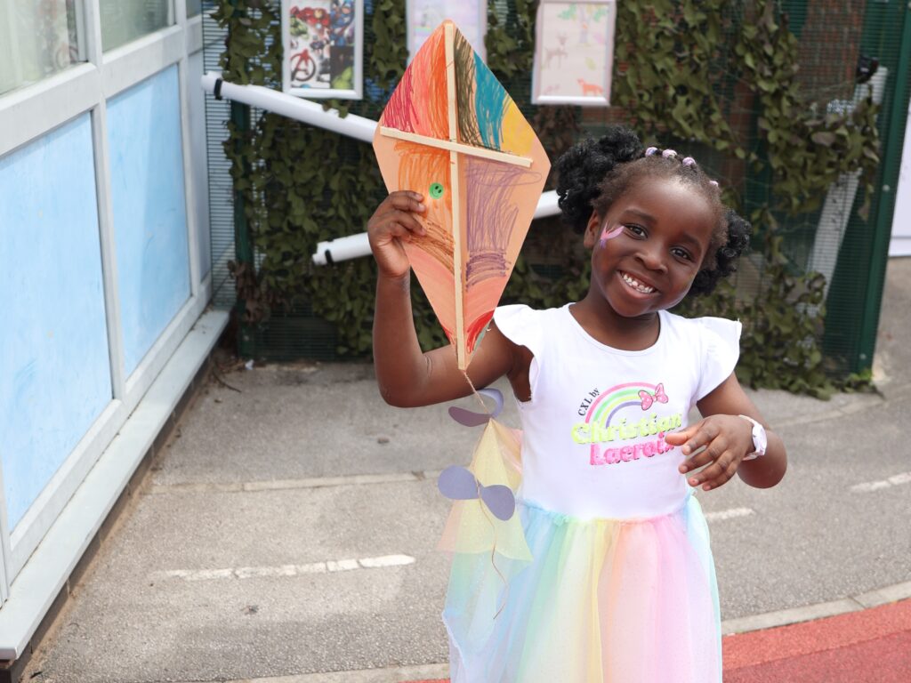 A little girl in a rainbow coloured dress holds up an orange kite
