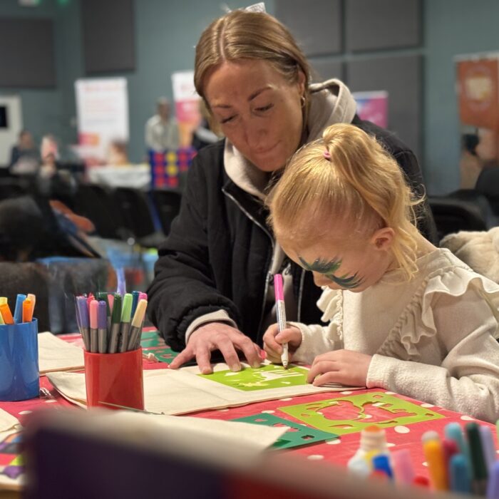 A mum sits next to her daughter, assisting her to draw