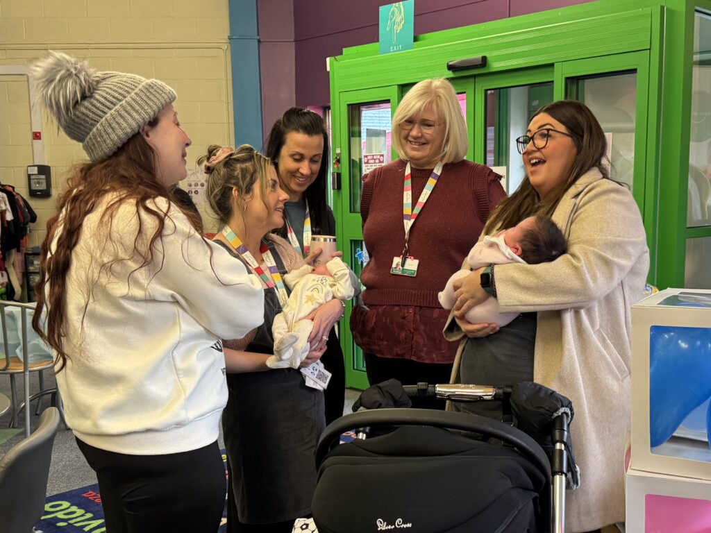 A group of mums holding babies stand in a circle chatting to Thrive at Five staff