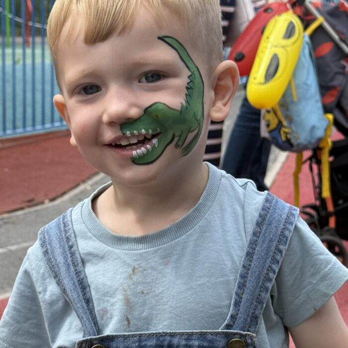 Boy wearing dungarees smiles at camera. His face is painted with a green dinosaur that curls around his mouth so that hie teeth appear to belong to the dinosaur.