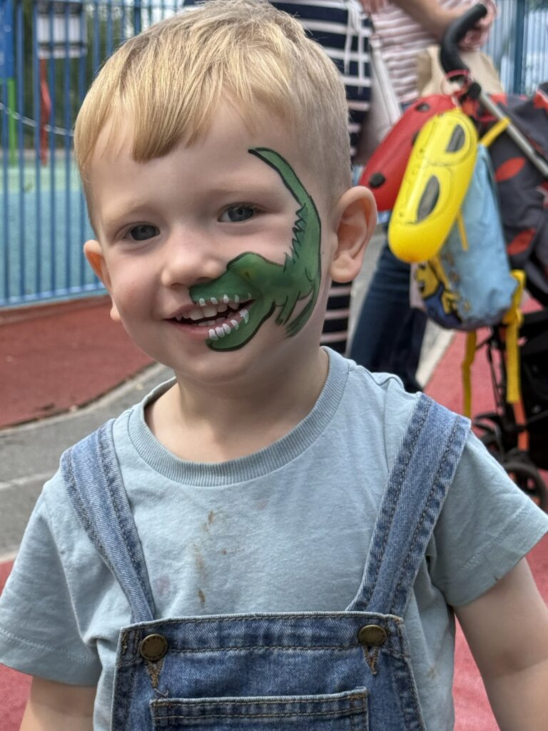 Boy wearing dungarees smiles at camera. His face is painted with a green dinosaur that curls around his mouth so that hie teeth appear to belong to the dinosaur.