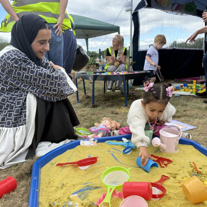 Mum in headscarf sits on grass by sandpit with daughter in white cardi and pink hair accessories, who is playing with a blue cup. In the background there are helpers and and a little boy in football kit.