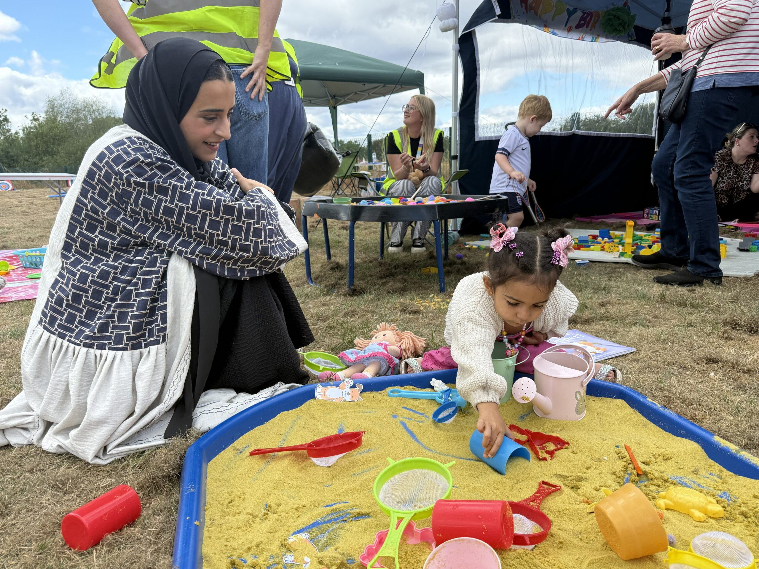 Mum in headscarf sits on grass by sandpit with daughter in white cardi and pink hair accessories, who is playing with a blue cup. In the background there are helpers and and a little boy in football kit.