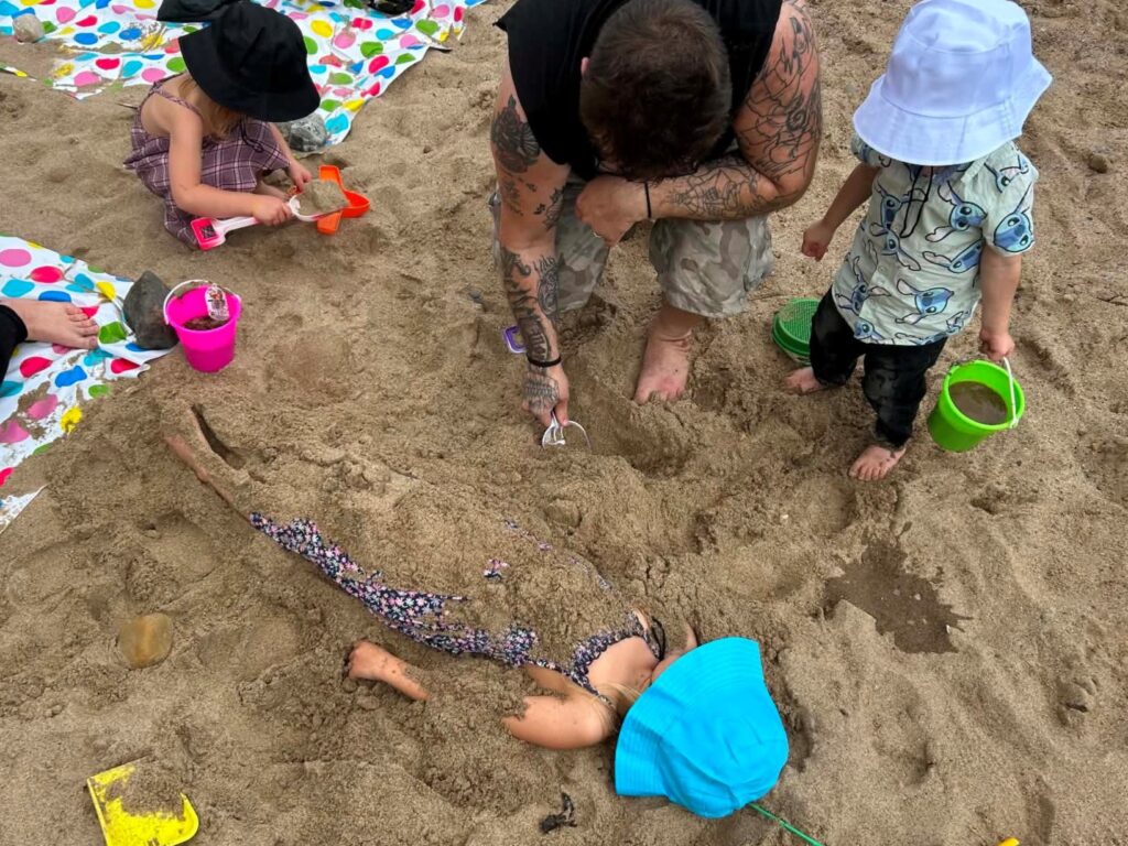 A child likes on the beach with most of their body covered in sand. Children and a male adult are nearby with buckets of sand to cover the child more.
