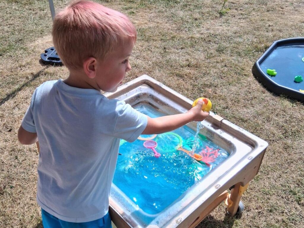 A boy stands at a table which has a big bowl of water and toys