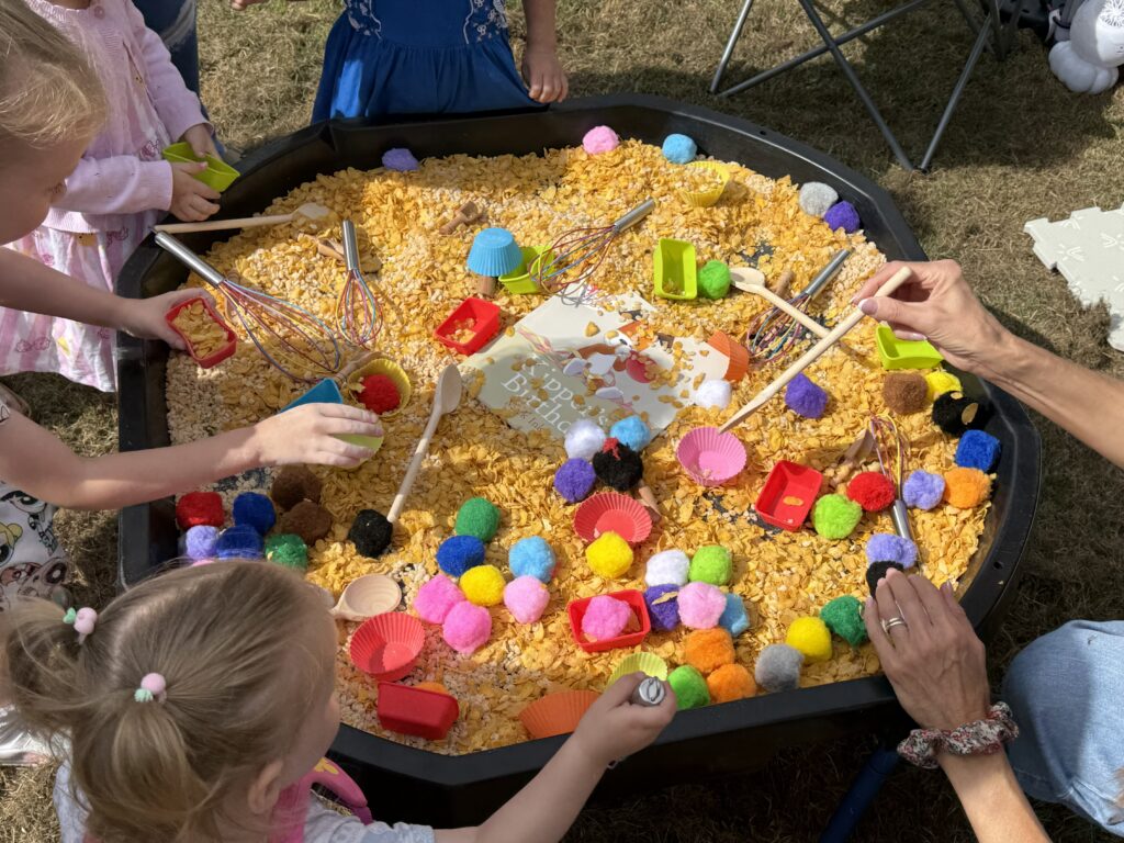 A tray full of cornflakes is surrounded by children as they play with spooning the cereal into containers