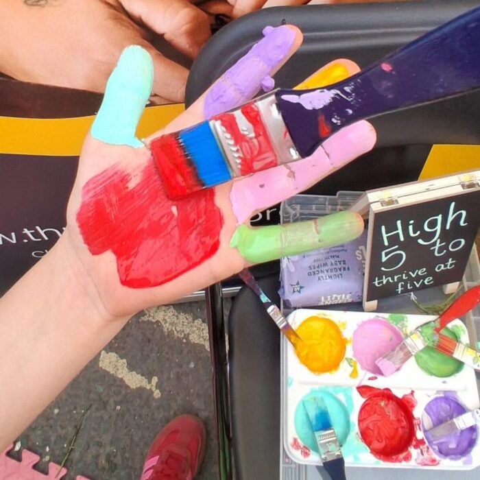 A child's hand is being painted with different colours on each finger to make a finger painting
