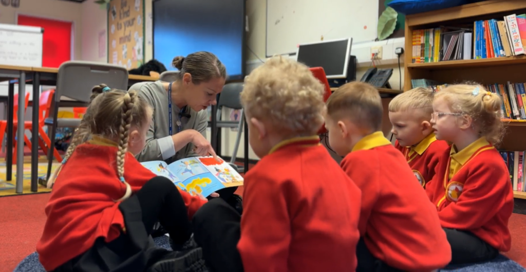 A group of nursery school children sit on the floor, gathered around the teacher as she points to pages in a book.