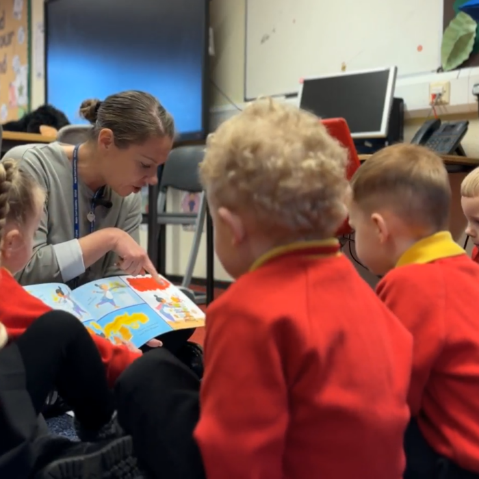 A group of nursery school children sit on the floor, gathered around the teacher as she points to pages in a book.